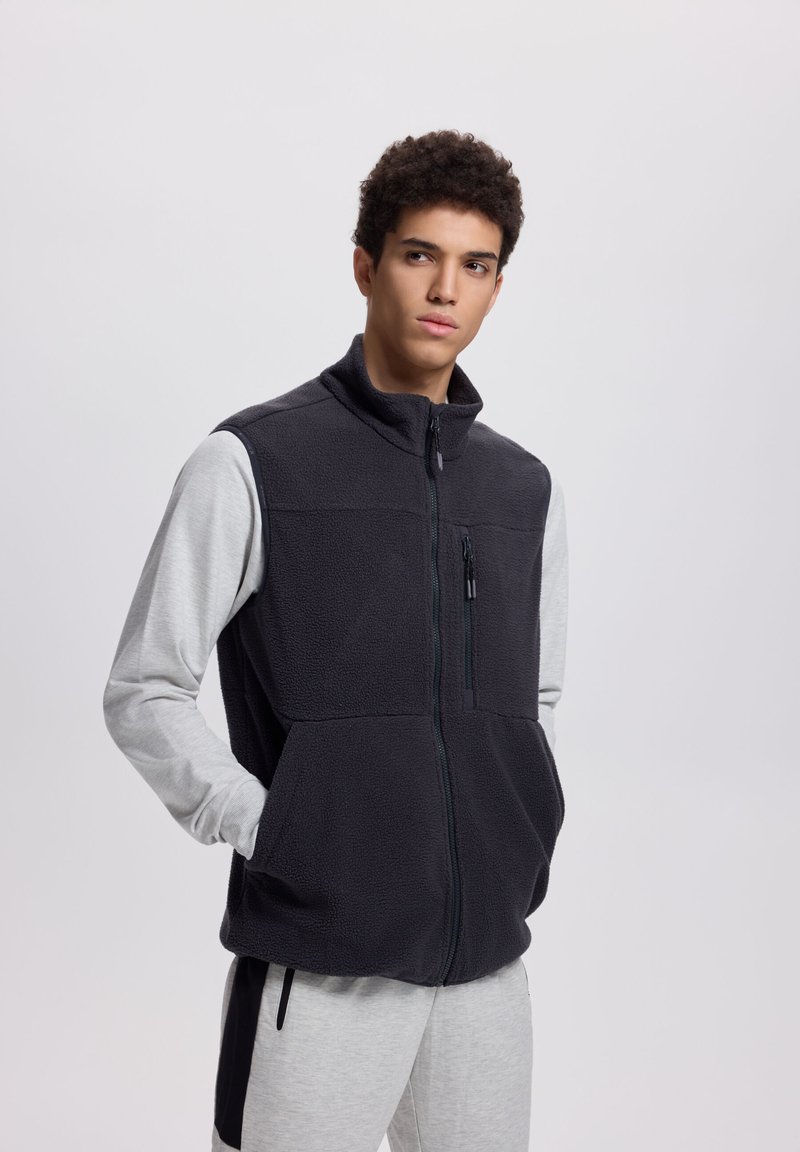 Giovane uomo che indossa un gilet in pile nero con cerniera sopra una maglia a maniche lunghe grigio chiaro e pantaloni della tuta coordinati, in piedi con le mani nelle tasche.