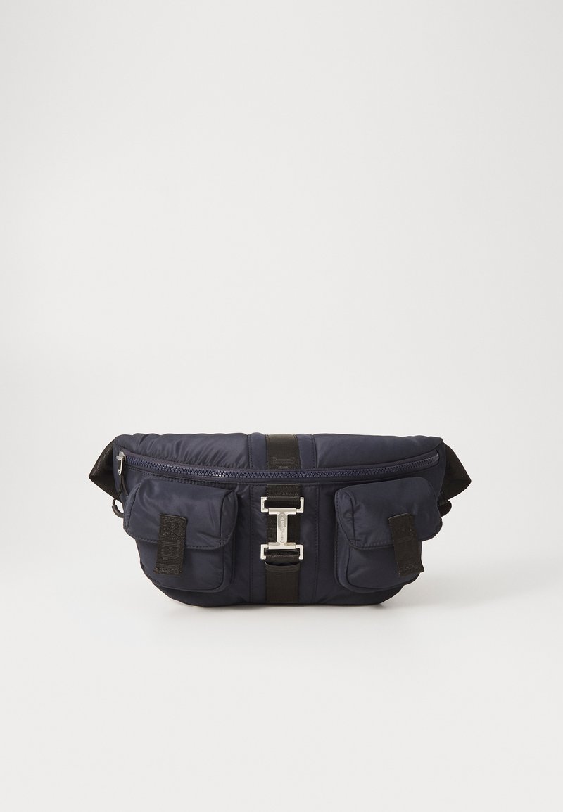 Navy nylon waist bag with a zip closure, two front pockets, black strap, and metallic hardware detail at the center. Smooth, padded texture.