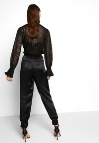 Svart genomskinlig blus med texturerat mönster och utsvängda ärmar, ihopparad med svarta satinjoggingbyxor med elastiska muddar och fickor.