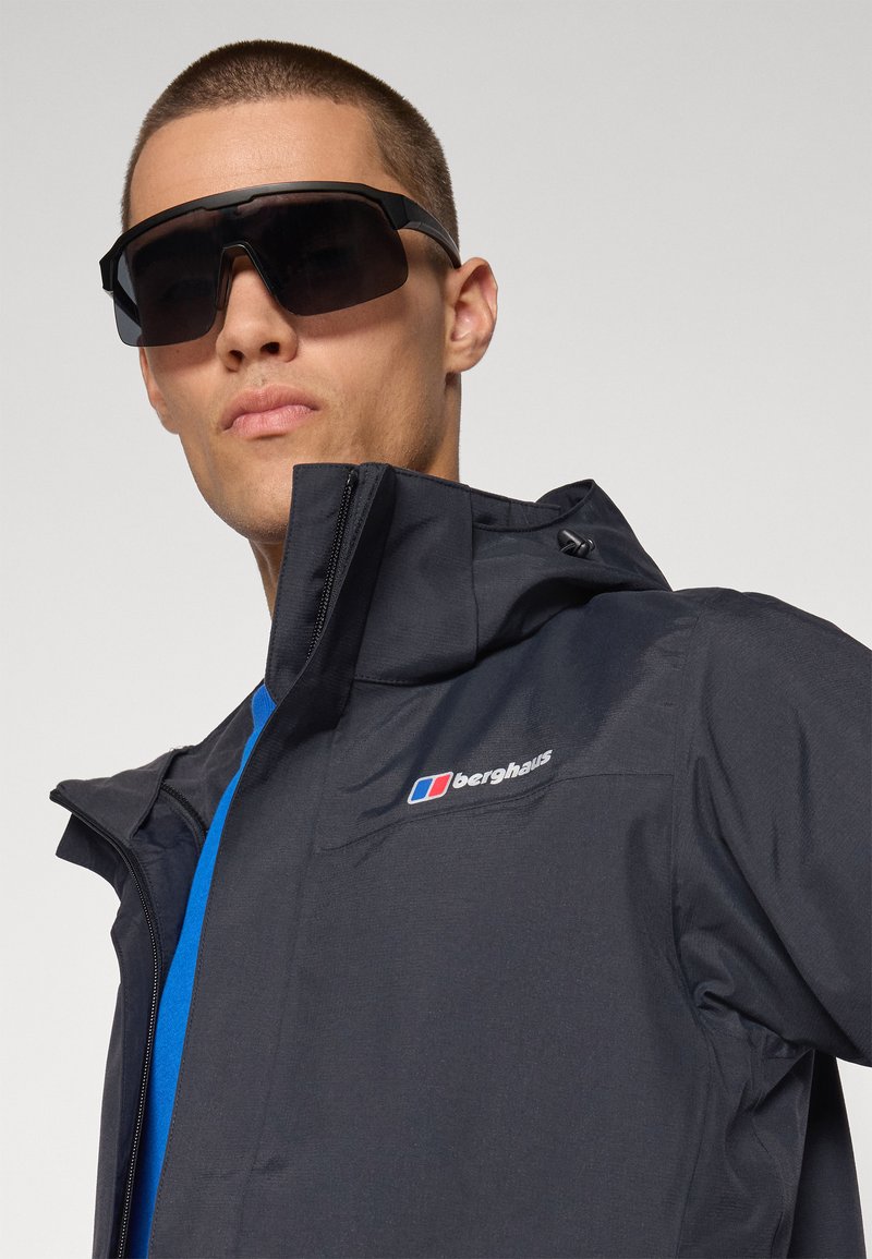 Czarna wodoodporna kurtka z kapturem, zapinana na zamek, z logo na lewej piersi. Nosi się ją na niebieskiej koszuli, w zestawie z dużymi ciemnymi okularami przeciwsłonecznymi.