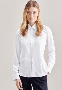 Camicia bianca a maniche lunghe con colletto classico, vestibilità slim e texture liscia, abbinata a pantaloni scuri. Senza fantasie né decorazioni.
