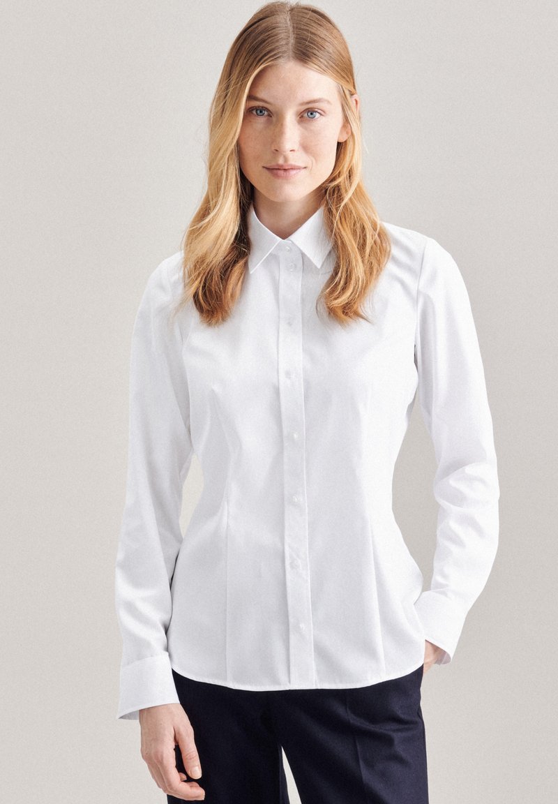 Camicia bianca a maniche lunghe con colletto classico, vestibilità slim e texture liscia, abbinata a pantaloni scuri. Senza fantasie né decorazioni.