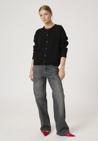 Schwarze Strickjacke mit strukturiertem Muster, runder Halsauschnitt, vordere Knopfleiste und zwei Vorderentaschen, kombiniert mit grauen weit geschnittenen Jeans.