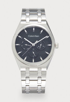 Montre en acier inoxydable argenté avec un cadran noir texturé de forme ronde. Comprend des index horaires blancs, un affichage de la date et trois sous-cadrans.