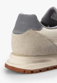 Zapatilla con un talón de ante gris texturizado y una parte superior suave color crema, que presenta una suela texturizada de color marrón claro y acentos de costura en gris.
