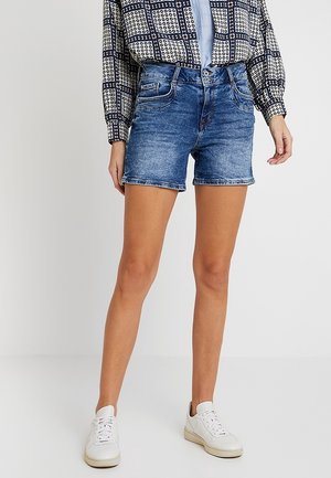 Kvinde iført blå denimshorts, en mønstret langærmet skjorte og hvide sneakers, stående foran en ensfarvet hvid baggrund.