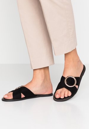 Sandales en suede noir à semelle plate avec des brides croisées et un accent métallique circulaire. Semelle plate avec des détails de couture visibles.