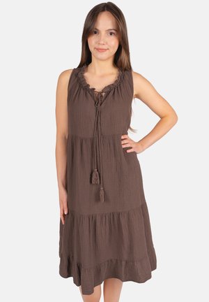 Femme debout, main sur la hanche, portant une robe marron sans manches à volants avec un col froncé et un lien à pompons.