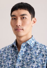 Wit overhemd met blauw paisleypatroon, knoopsluiting onder de kraag en gestructureerde stof. Close-up toont gedetailleerd ontwerp en kleuren.