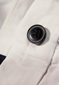 Bouton noir à quatre trous cousu sur un tissu blanc avec une couture visible et des plis dans le matériau.