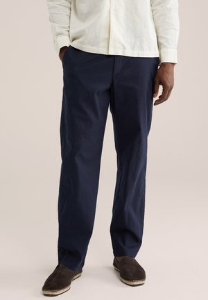 Muž nosiaci tmavomodré neformálne nohavice, svetlobéžovú košeľu s dlhým rukávom a tmavohnedé nazúvacie topánky, stojaci s jednou rukou v vrecku.