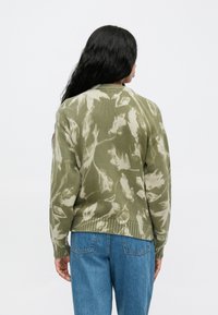 Žalias megztinis su grietinėlės spalvos tinklelio raštu, su įpjovomis apykakle ir rankovėmis, laisvas stilius, derinamas su mėlynomis džinsinėmis kelnėmis.