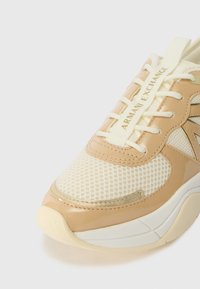 Beige és krém színű sneaker, hálós anyaggal, fényes részletekkel és arany díszítéssel. Jellemzője a texturált minta és a márkázott fűző címke.