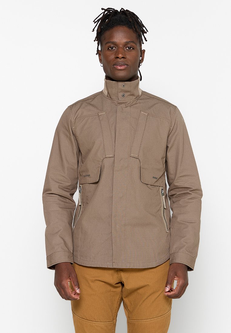 Chaqueta marrón con un cuello alto, dos bolsillos frontales con solapas y una textura suave. Costuras laterales con cremallera y sutiles detalles de costura.