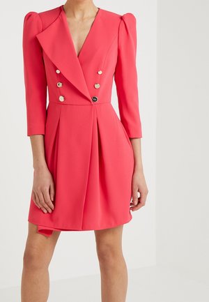 Kobieta ubrana w koralowo-różową sukienkę żakietową na dwa rzędy z złotymi guzikami i półdługimi bufkami na rękawach, stojąca na tle jednolitego białego tła.