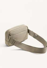 Borsa crossbody in tessuto beige con forma rettangolare, bordi arrotondati, tasca con zip e tracolla regolabile. Superficie testurizzata.