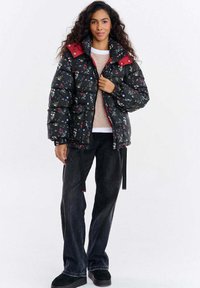 Czarna pikowana kurtka w kwiatowy wzór z czerwonymi akcentami i wysokim kołnierzem, noszona na jasnym swetrze, zestawiona z ciemnymi dżinsami i czarnymi butami.