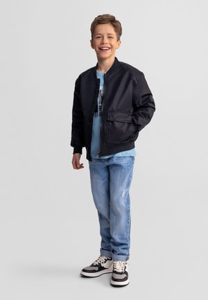 Uśmiechnięty chłopiec stojący, ubrany w czarną kurtkę, niebieski t-shirt z grafiką, jasnoniebieskie dżinsy i czarno-białe trampki na jednolitym tle.