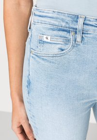 Vaqueros de mezclilla de color azul claro con un diseño de cintura alta, cinco bolsillos y una etiqueta de logo en el bolsillo trasero. Textura suave con un ligero desgastado.