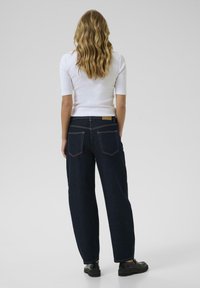 Vaqueros de pernera ancha de un azul oscuro con una cintura alta, bolsillos traseros visibles y costuras en contraste. Combinados con una camiseta ajustada blanca y zapatos negros.