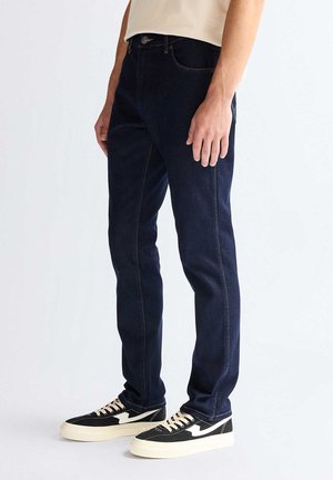 Persona che indossa jeans slim fit blu scuro e sneakers nere e bianche, in piedi davanti a uno sfondo chiaro e uniforme.