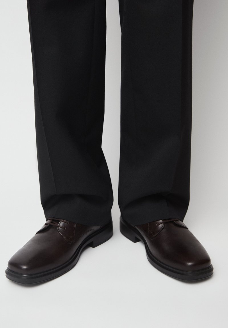 Dunkelbraune Lederschuhe, getragen mit schwarzen formellen Hosen auf einem schlichten hellen Hintergrund.