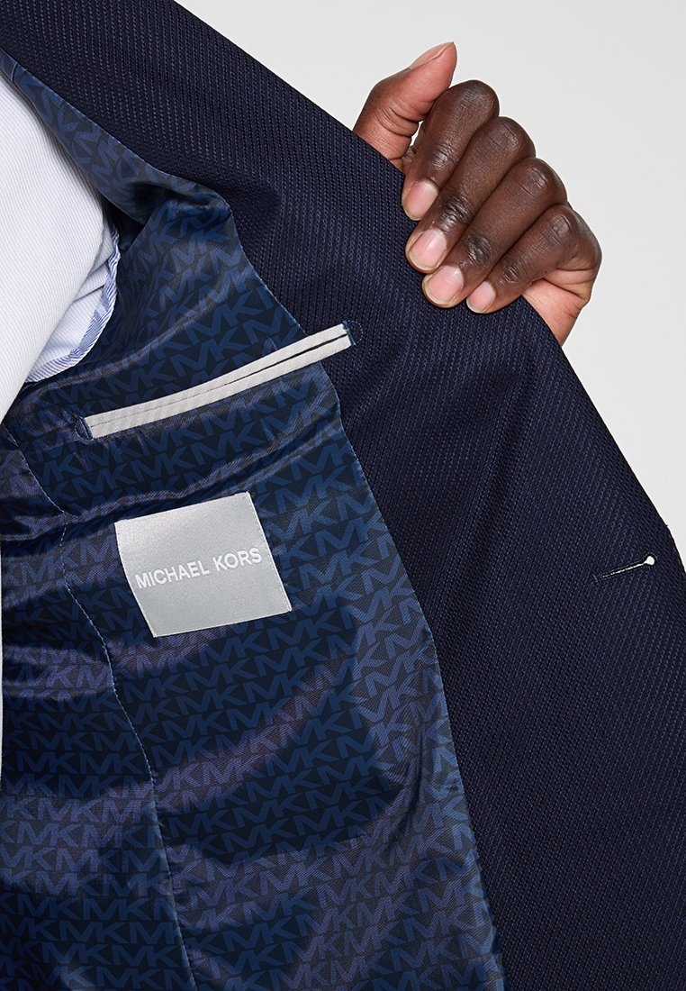 Innenansicht eines marineblauen Blazers mit einem strukturierten Muster, ausgestattet mit einem glatten Satinfutter und einem Etikett, das "MICHAEL KORS" liest. Enthält eine Innentasche.