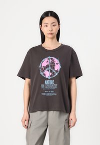Afslappet brun t-shirt med et globe-grafik i pink og blå, tekst med teksten "NATURE VS INDUSTRY," kortærmet og med rund hals.