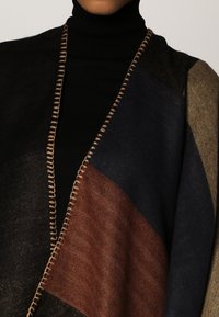 Black turtleneck under a color-blocked wool shawl with brown, navy, beige sections and decorative stitching along the edge.