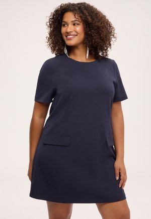 Frau mit lockigem Haar, die ein marineblaues Kleid mit kurzen Ärmeln und zwei vorderen Taschen trägt, lächelnd und seitlich schauend.