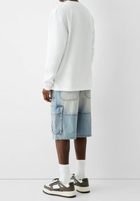 Bílá košile s dlouhým rukávem, modré denimové cargo šortky s velkými kapsami a šedé tenisky. Textura vypadá měkce a materiály jsou ležérní.