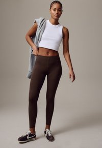Braune Leggings, kombiniert mit einem weißen, bauchfreien Tanktop. Ein hellgrauer Pullover ist über eine Schulter gelegt. Schwarze und weiße Turnschuhe runden den Look ab.