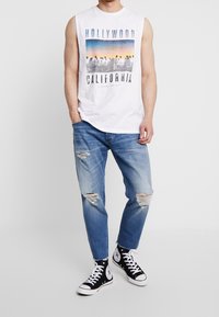 Vit ärmlös grafisk t-shirt med trycket "HOLLYWOOD CALIFORNIA", kombinerad med ljusblå slitna jeans och svarta högstövlar.