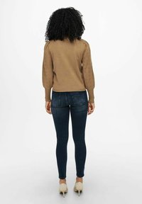 Pull marron en maille avec manches ballon et détails œillets, associé à un jean skinny foncé et des chaussures à talons beiges.