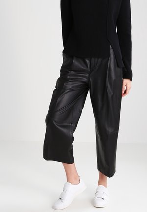 Person iført sorte læderbukser, sorte langærmede top og hvide sneakers med velcrolukning, stående mod en ensfarvet baggrund.