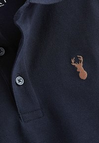 Marineblauwe poloshirt van gestructureerde stof, met een klein bruin geborduurd hertlogo en twee grijze knopen op de kraag.