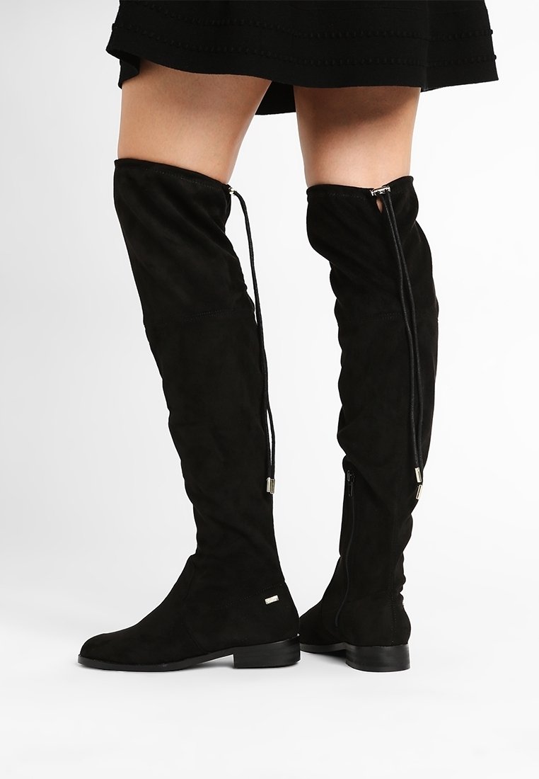 Bottes en daim noir au-dessus du genou avec talons bas, fermetures éclair latérales et cordons décoratifs portées avec une jupe noire sur fond blanc.