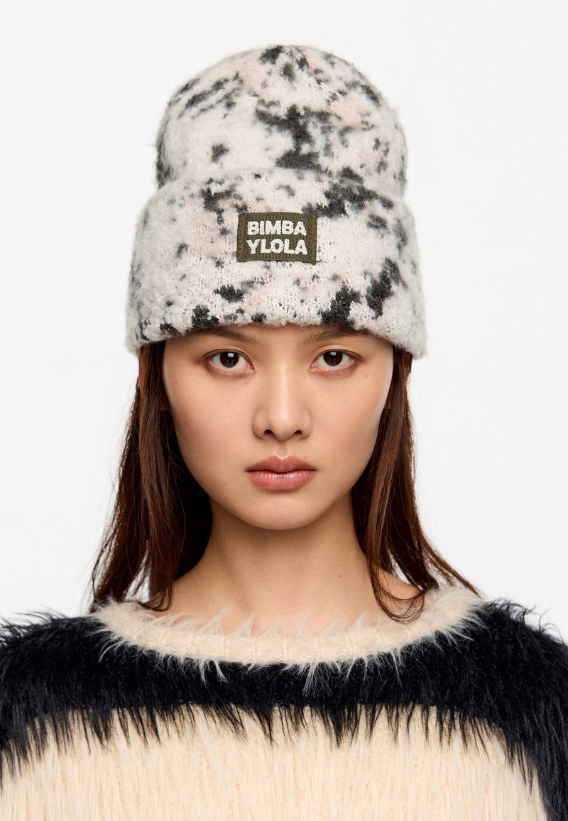 Kootud beanie valge värvusega musta ja roosa marmoriga, millel on roheline kangasääre silt "BIMBA YLOLA". Pehme, tekstuuriga materjal.
