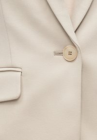 Blazer beige clair avec col cranté, tissu texturé, poche latérale et bouton arrondi à quatre trous pour la fermeture.