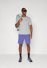 Gray short-sleeve t-shirt with blue logo, paired with purple shorts. Carrying a light blue backpack and wearing black sunglasses.