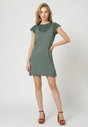 Mujer joven con top floral verde debajo de un vestido sin mangas verde salvia con sandalias marrones de plataforma, de pie con la mano en la cadera.