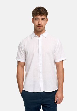 Mann mit kurzen lockigen Haaren, der ein weißes kurzärmeliges Hemd mit Knöpfen und dunkle Hose trägt, steht vor einem einfarbigen Hintergrund.