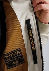 Lightweight bomber jacket with a cream outer fabric and yellow mesh lining. Features black fabric labels with gold text and a zipper closure.