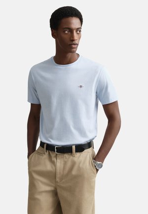 Hellblaues kurzärmeliges T-Shirt mit rundem Ausschnitt und kleinem gesticktem Detail auf der Brust, kombiniert mit beigen Hosen und einem schwarzen Gürtel.