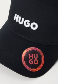 Svart caps laget av teksturert stoff, med hvit brodert "HUGO" foran og et rundt merke klistremerke på visiret.