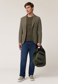 Lichte olijfgroene blazer met bruine knopen, gecombineerd met een donkergroene shirt en blauwe spijkerbroek. Draagt een donkergroene rugzak met zwarte accenten.