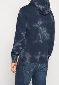 Tmavě modrá mikina s kapucí a batikovacím vzorem, měkký materiál, kangaroo kapsa, šňůrka na kapuci a žebrované manžety. Kombinováno s tmavě modrými džínami.