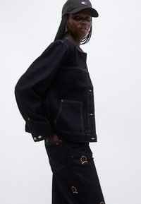 Γυναίκα με σκούρο τζιν μπουφάν και παντελόνι με κεντημένα σήματα, φορώντας μαύρο καπέλο Tommy Jeans, με τα χέρια στις τσέπες, σε λευκό φόντο.