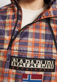 Skakajakke i orange, lilla og beige med frontlynlås og "Napapijri geographic" logo patch over et norsk flag patch.