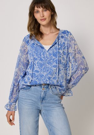 Kobieta nosząca przezroczystą niebieską bluzkę w kwiatowy wzór z długimi rękawami oraz jasnoniebieskie, wysokie jeansy, stojąca z jedną ręką w kieszeni.